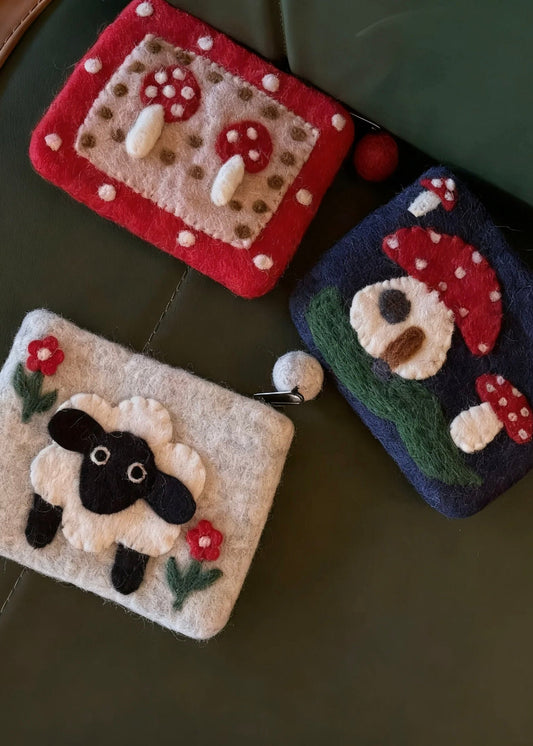 A collection of three handmade wool felt coin purses featuring a black sheep, red mushroom dots, and a forest mushroom house design.